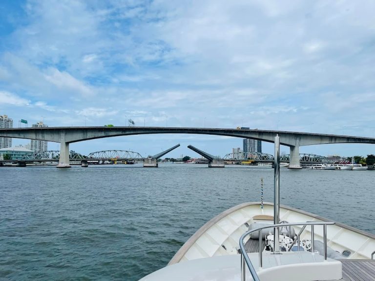 Superyachts Krungthep Bridge: Bangkok's blue Chao Phraya river is seen from the bow of a superyacht below a blue sky with clouds forming, the krungthep bridge is open in the near distance.