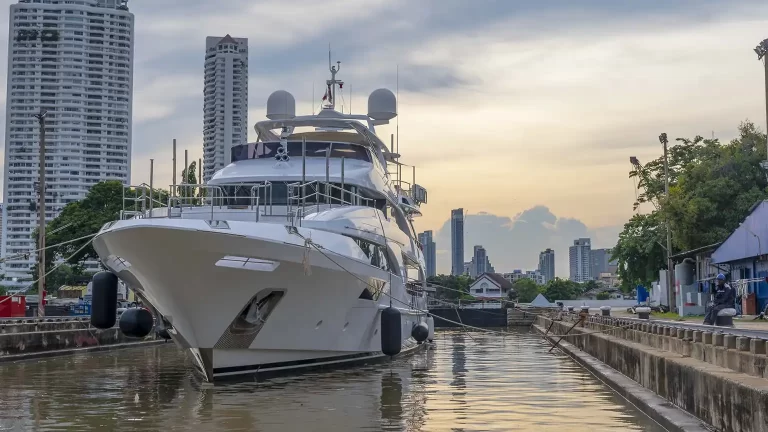 The large white M/Y Asia Poseidon is docked in a dock, tall modern buildings and trees under a cloudy evening sky, awaiting a yacht refit in the bangkok dry docks