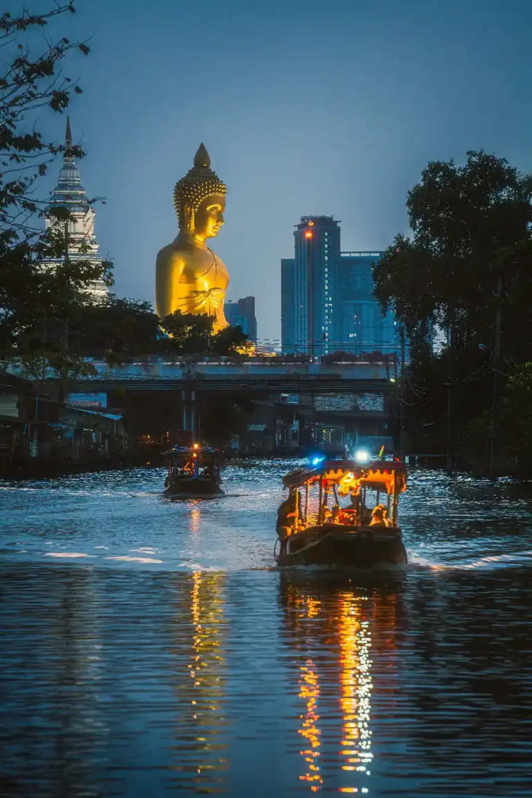Evening boat ride on a Bangkok canal with the illuminated Great Buddha of Wat Paknam towering over the skyline, showing the contrast of traditional Thai culture and modern city life.